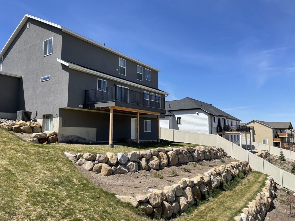 A house with a stone wall and a fence, featuring a design by Crown Finish, set against a clear blue sky.