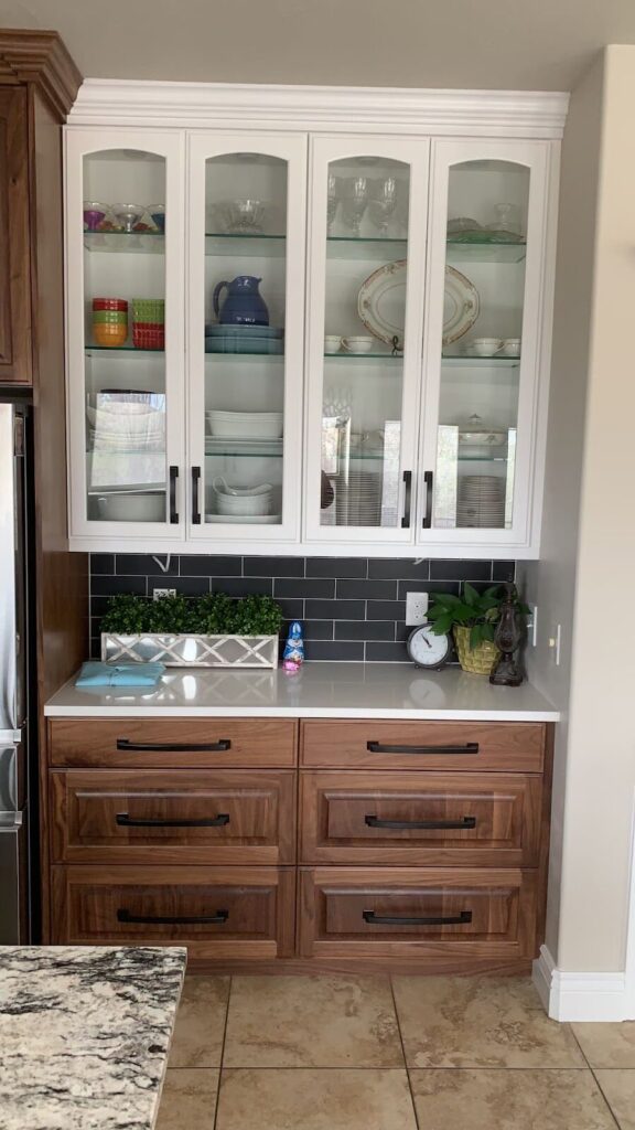 A modern kitchen with white glass-front cabinets, wooden drawers, black subway tile backsplash, and a marble countertop featuring plants and decor.