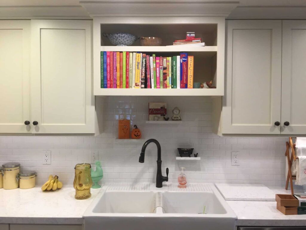 White kitchen with farmhouse sink, black faucet, and colorful cookbooks on open shelving.