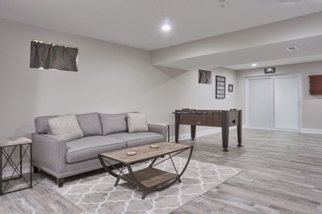 A spacious, modern finished basement featuring a grey fabric sofa, a geometric rug, a wooden coffee table, and a foosball table on grey wood-plank flooring.